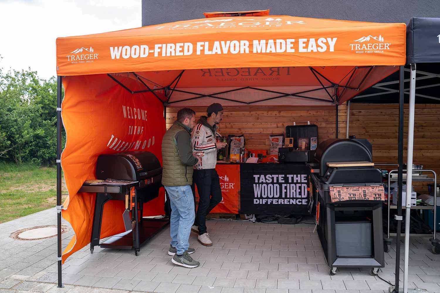 Two people stand at a wood-fired grill display under an orange tent.