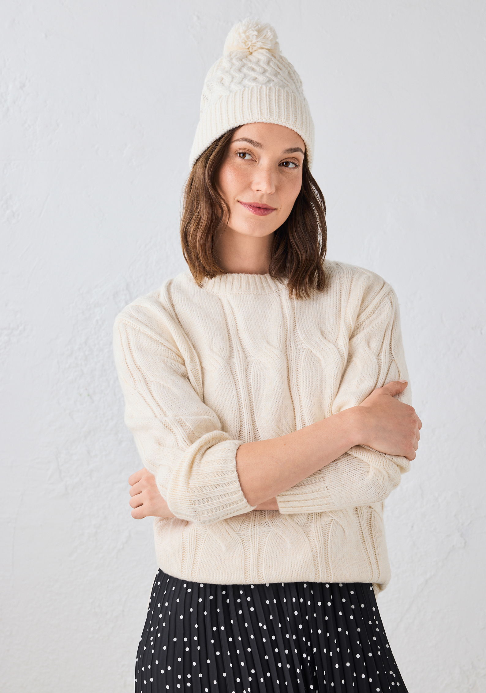 Young woman wearing a white cable-knit sweater and matching beanie, arms crossed.