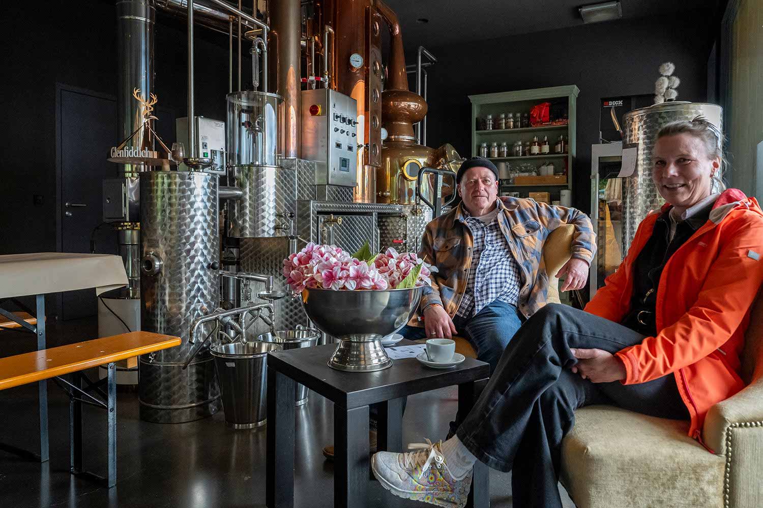 Two men sitting in a distillery, surrounded by equipment and decor.