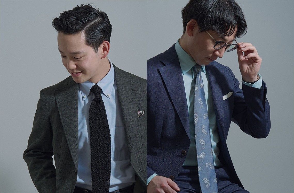 Two men in suits, one adjusting glasses, the other smiling.