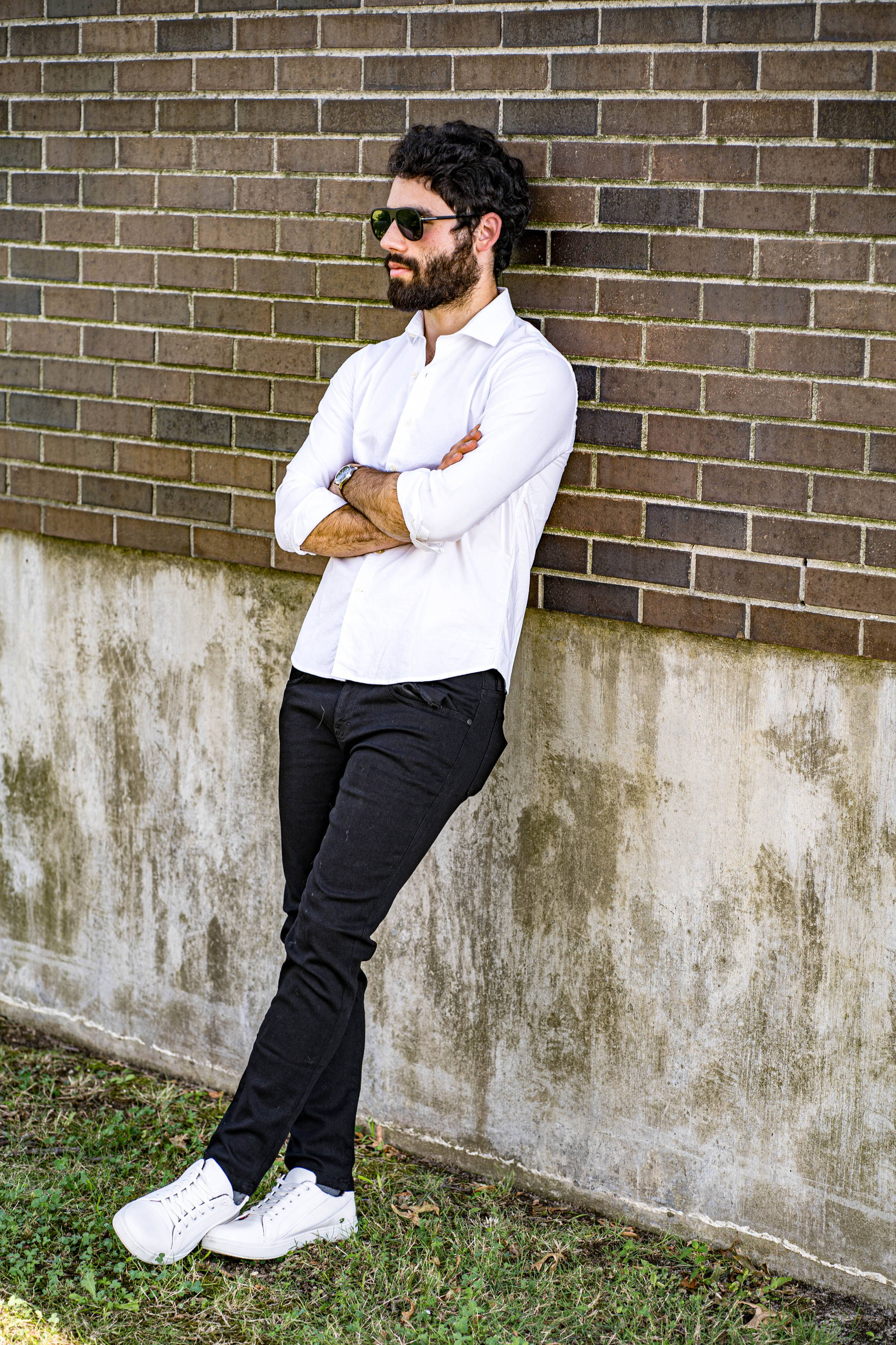 Man leaning against brick wall folded arms wearing a white button down shirt, white sneakers, and black jeans for short men from under510.com