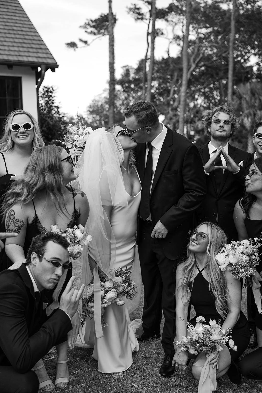 Classic black and white photograph capturing a heartwarming moment as the bride lovingly kisses the groom, while surrounded by joyful bridesmaids.