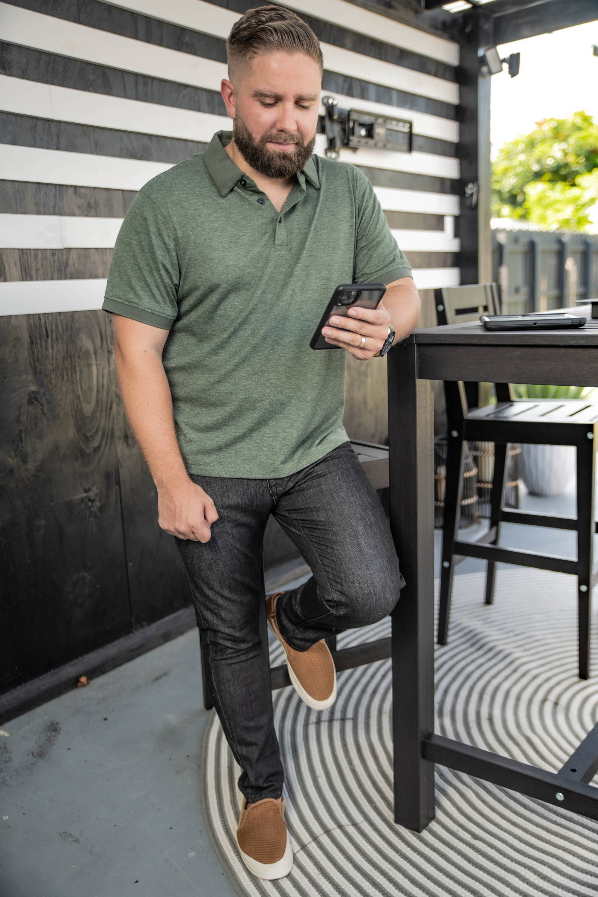 Man looking at phone wearing brown shoes, green polo shirt, and black stretch jeans from under510.com