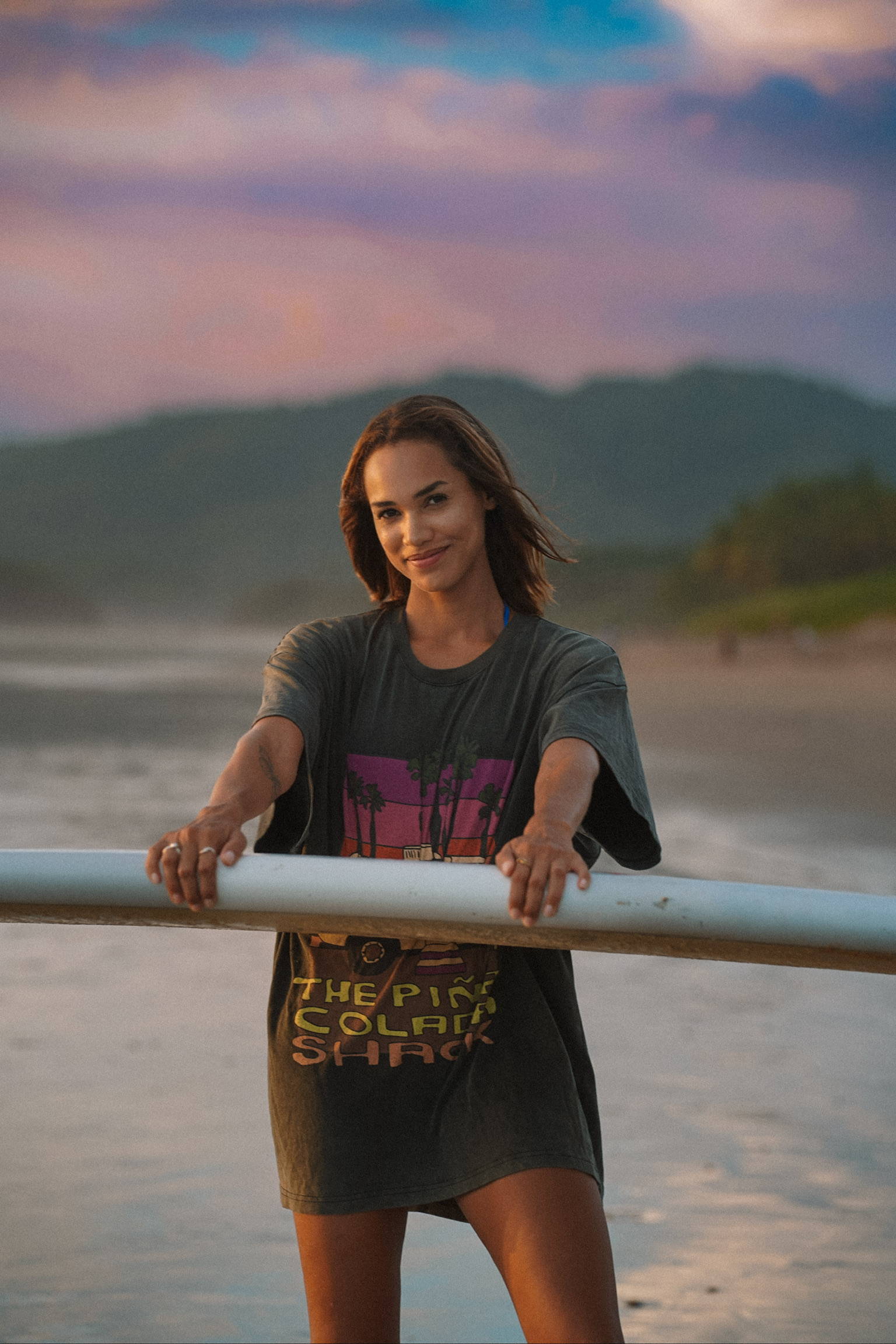 A woman wearing an oversized graphic t-shirt holding her surfboard on a beach in Costa Rica