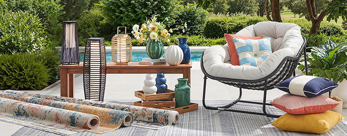 Outdoor seating area with decorative pillows, rugs, and flowers by a pool.