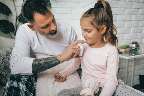Ein Vater hilft seiner Tochter bei der Anwendung eines Nasensprays, das ein Antihistaminikum enthält, um ihre durch Heuschnupfen verstopfte Nase wieder zu befreien.