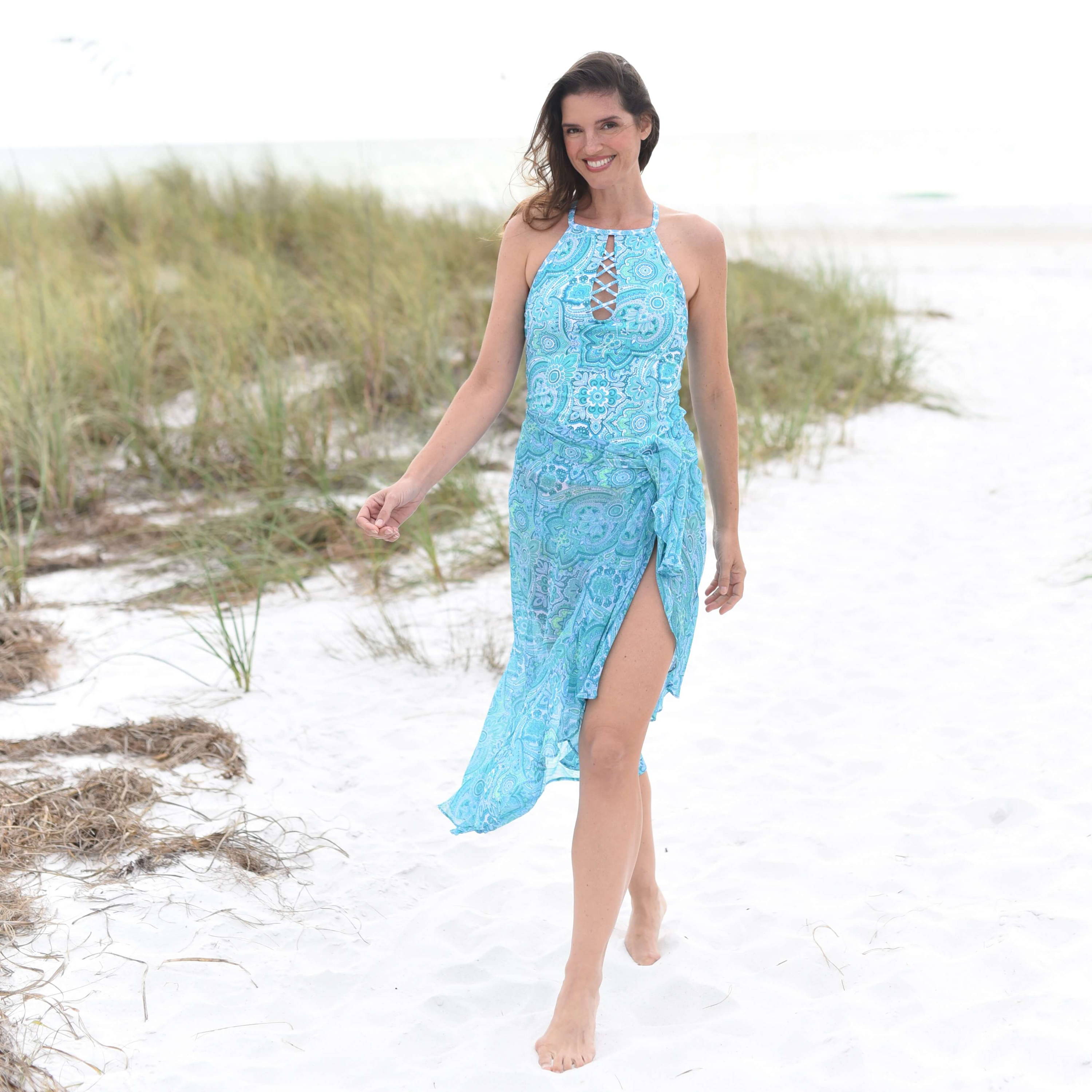 A smiling brunette woman wearing a blue bathing suit and sarong walks across the sand at the beach