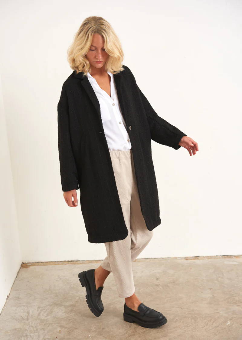 A model wearing a black, midi cocoon coat over a white shirt, off white trousers and black leather loafers