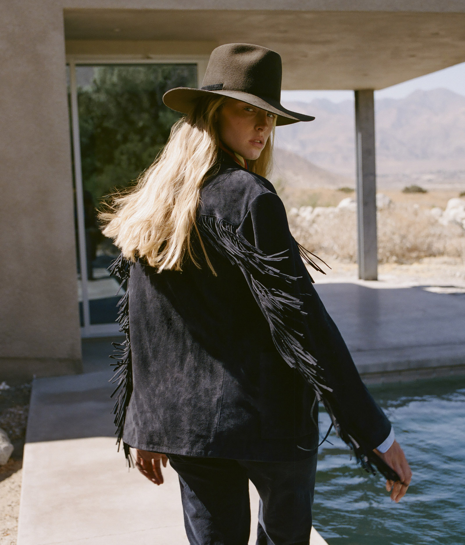 Woman in a fringed jacket and hat, standing by a pool.