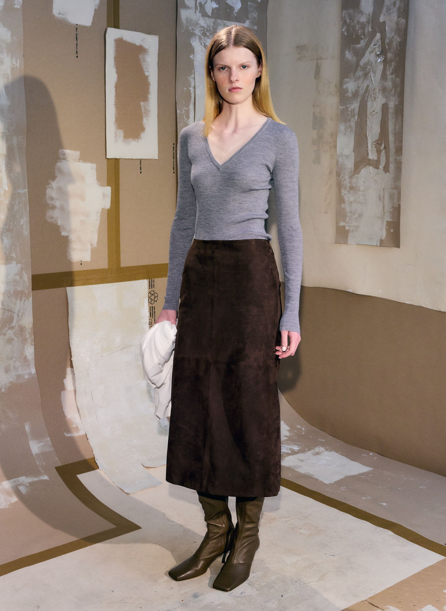 Model wearing a gray top and brown skirt, posing against an artistic backdrop.