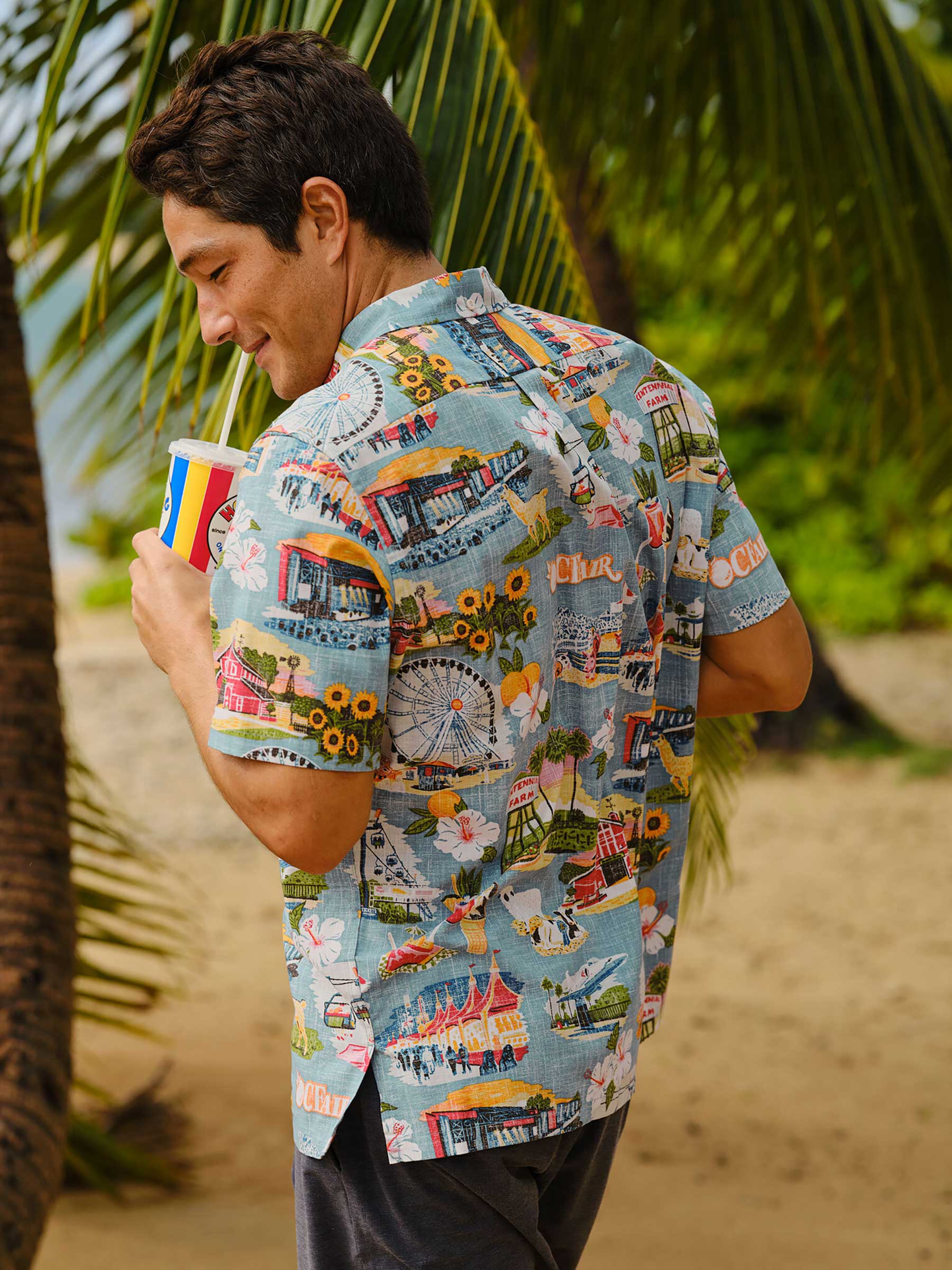 Man in a colorful shirt holding a drink, standing by palm trees on a beach.