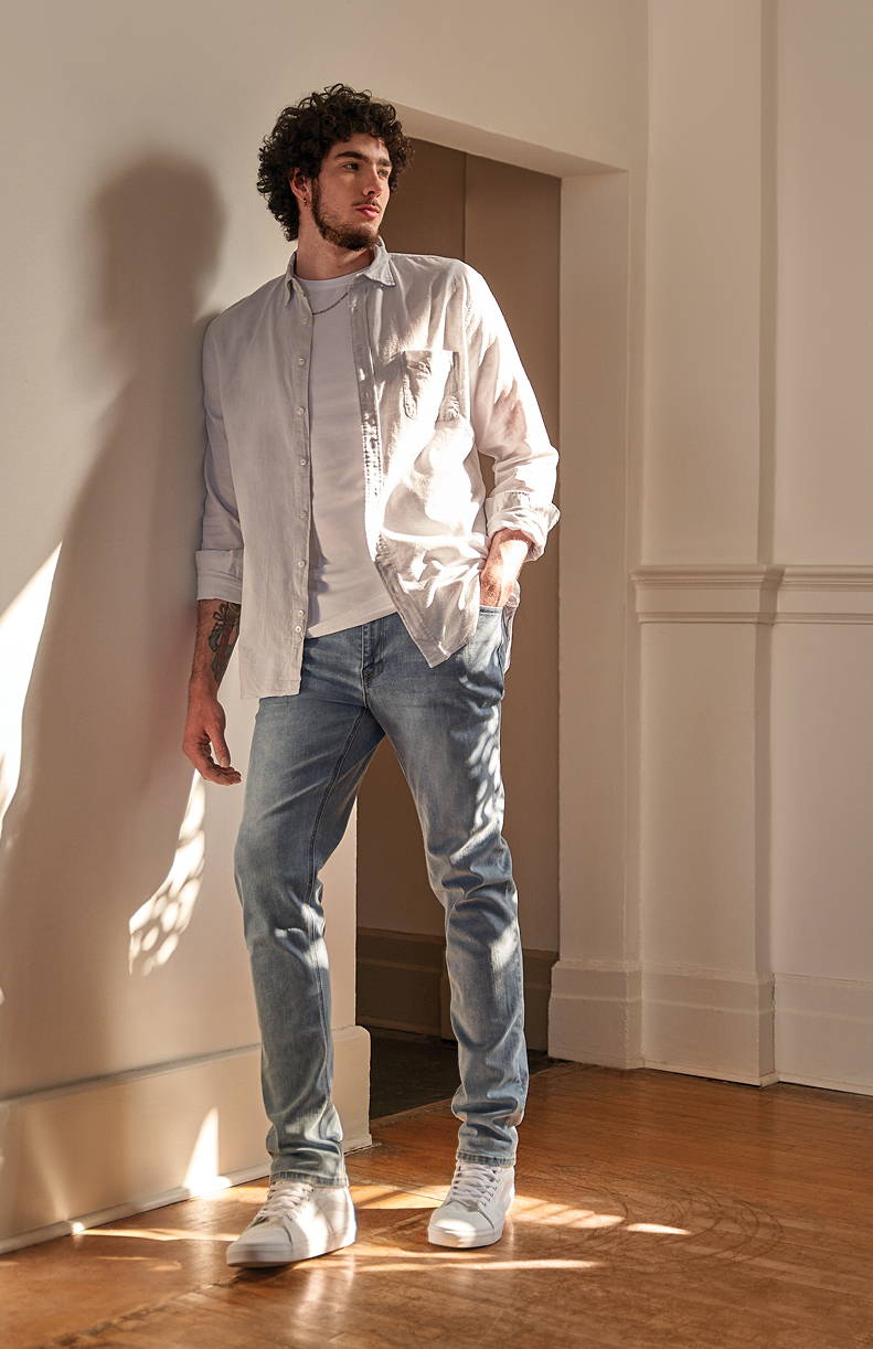 Tall man leaning against a wall wearing a white linen shirt and light blue slim jeans.