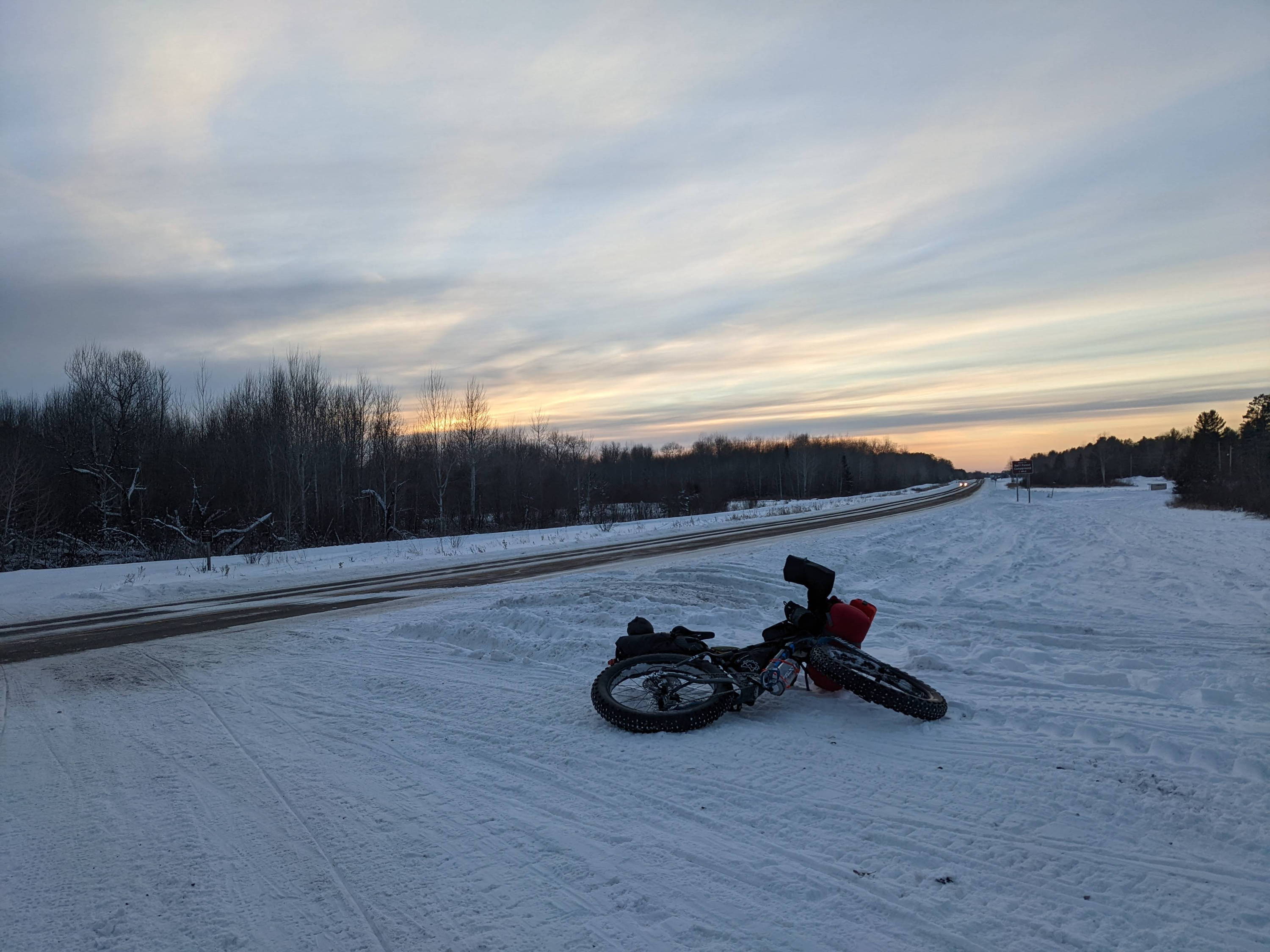 A fully-loaded bikepacking bike in the snow by a paved road with a cloudy sunset in view.