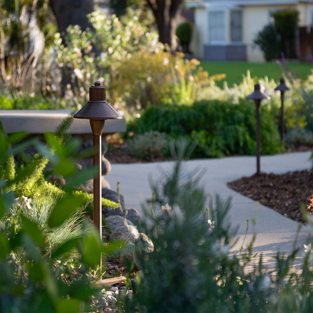 A concrete path with landscaping brush and copper pathway lights.