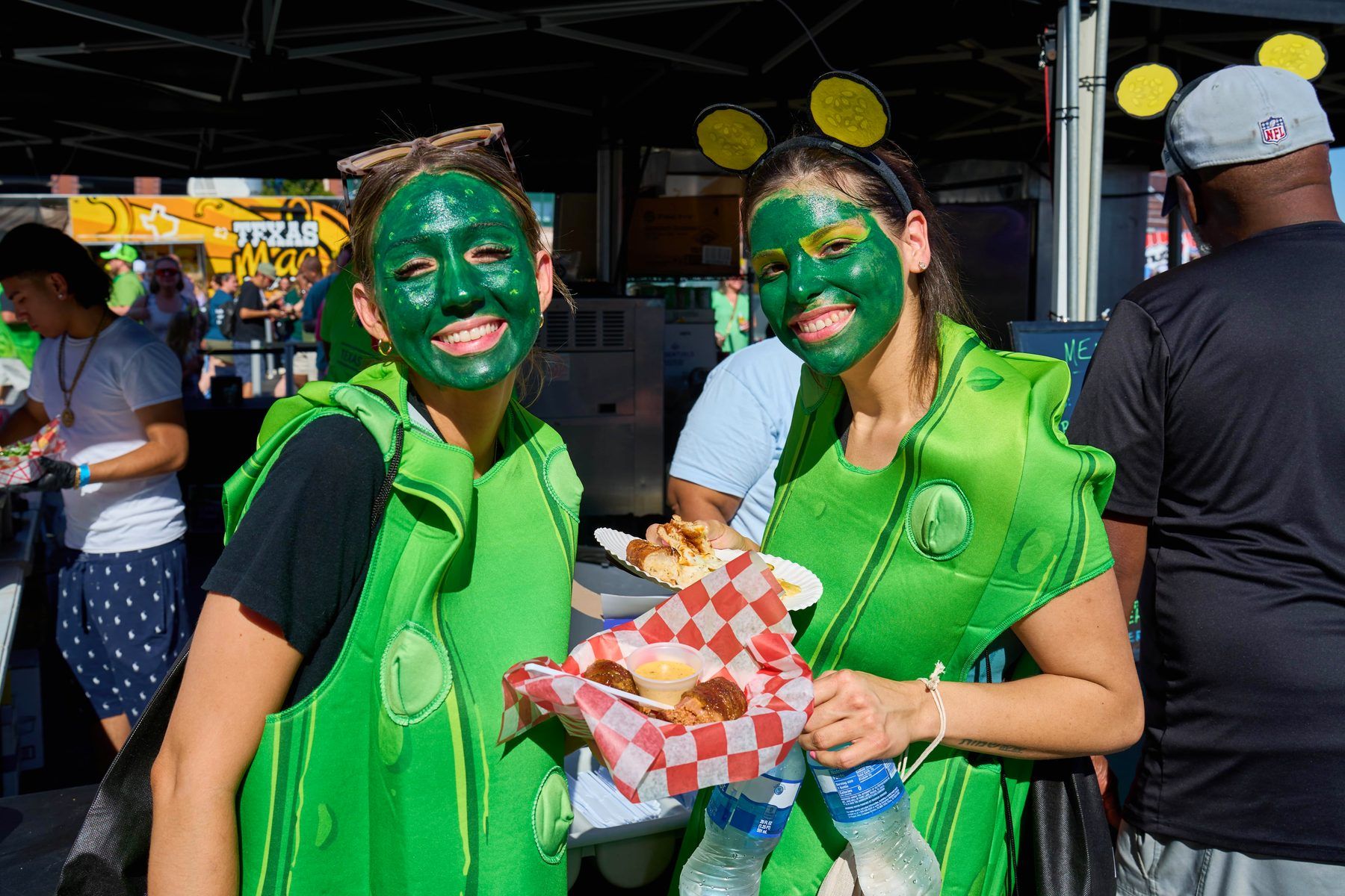 Influencers in pickle costumes with full green face paint and pickle slice headbands holding pickle festival food at The Big Dill Arlington Texas 2025.