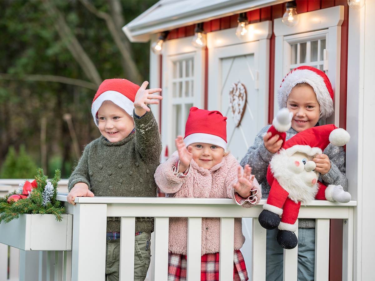 Kids waving on the porch of  the red and white wooden playhouse with Christmas decorations by WholeWoodPlayhouses