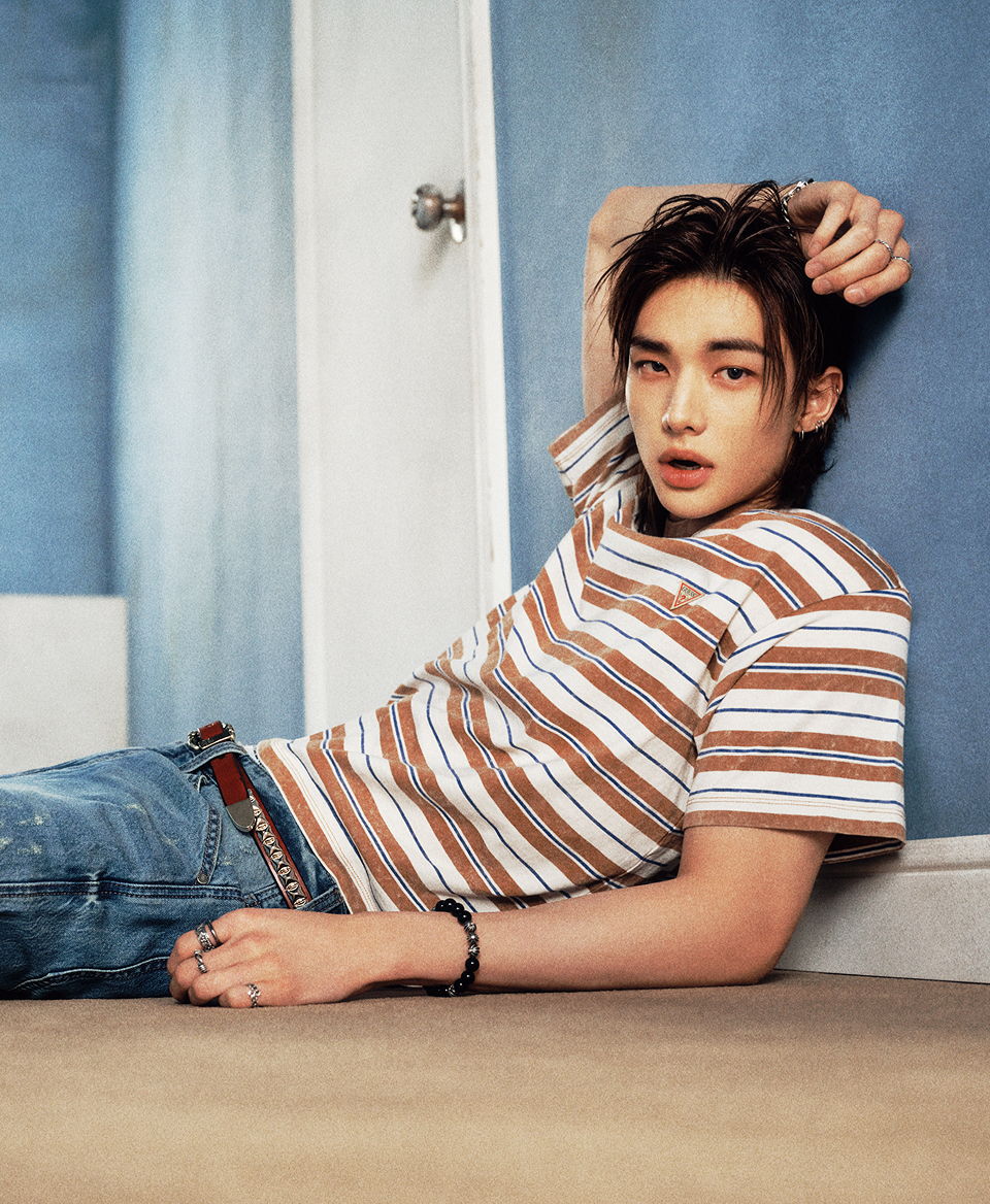 Hyunjin lounging against a wall, wearing a striped shirt and jeans.