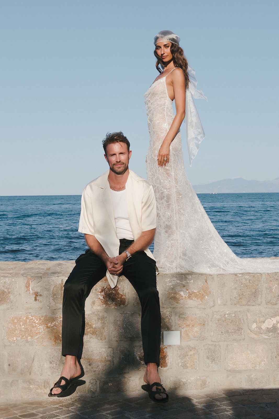 A man sitting on a wall with a woman in a wedding dress by the sea.