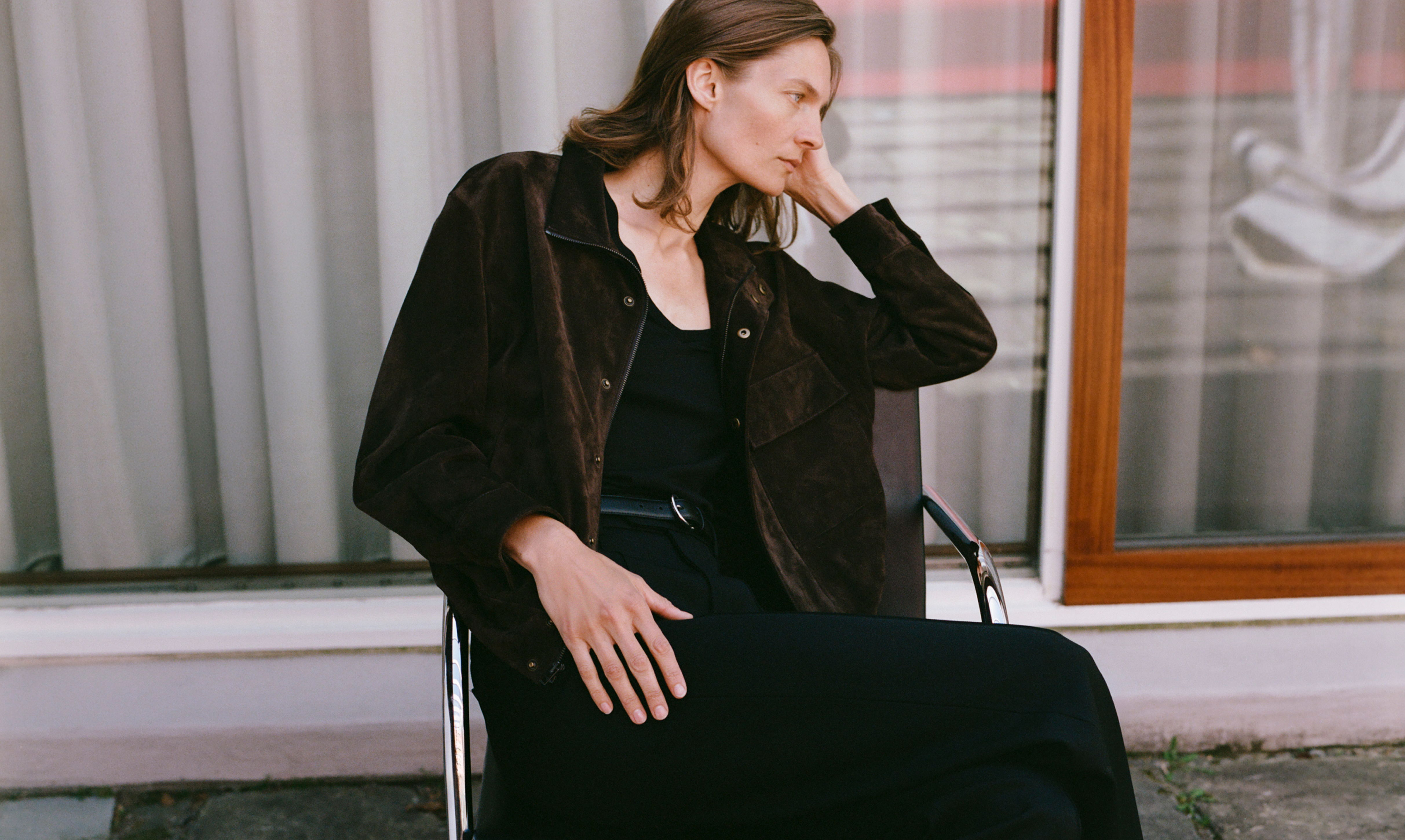 Model sitting in a chair, wearing a black dress and brown jacket.