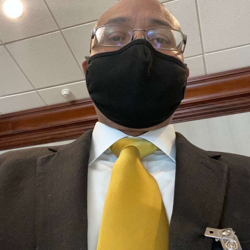 Man taking a selfie wearing a black suit and gold solid tie as part of a work uniform