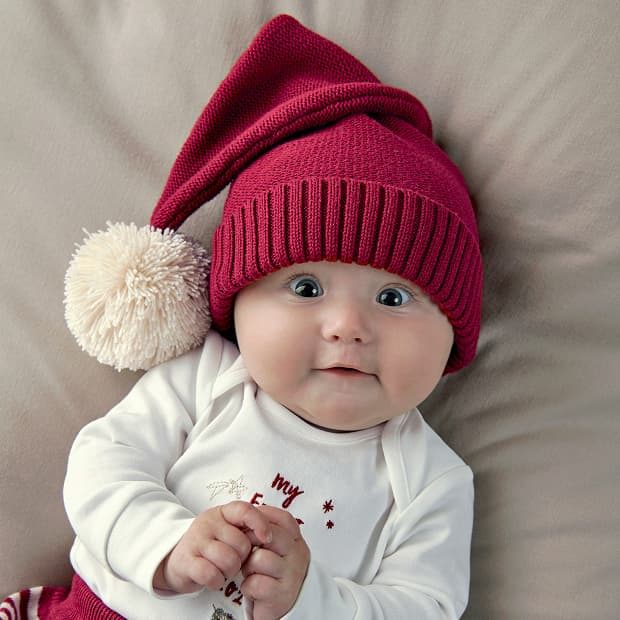 Baby wearing a red knitted hat with a pom-pom, smiling on a soft surface.