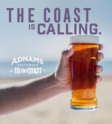 A hand holding a glass of beer on a beach.