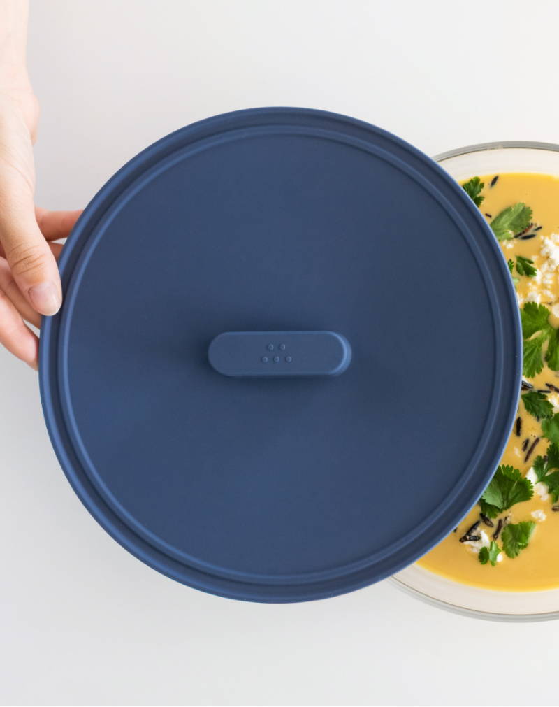 A hand removes a Blue Misen Universal lid from a glass container holding yellow soup topped with parsley, cheese, and pepper.
