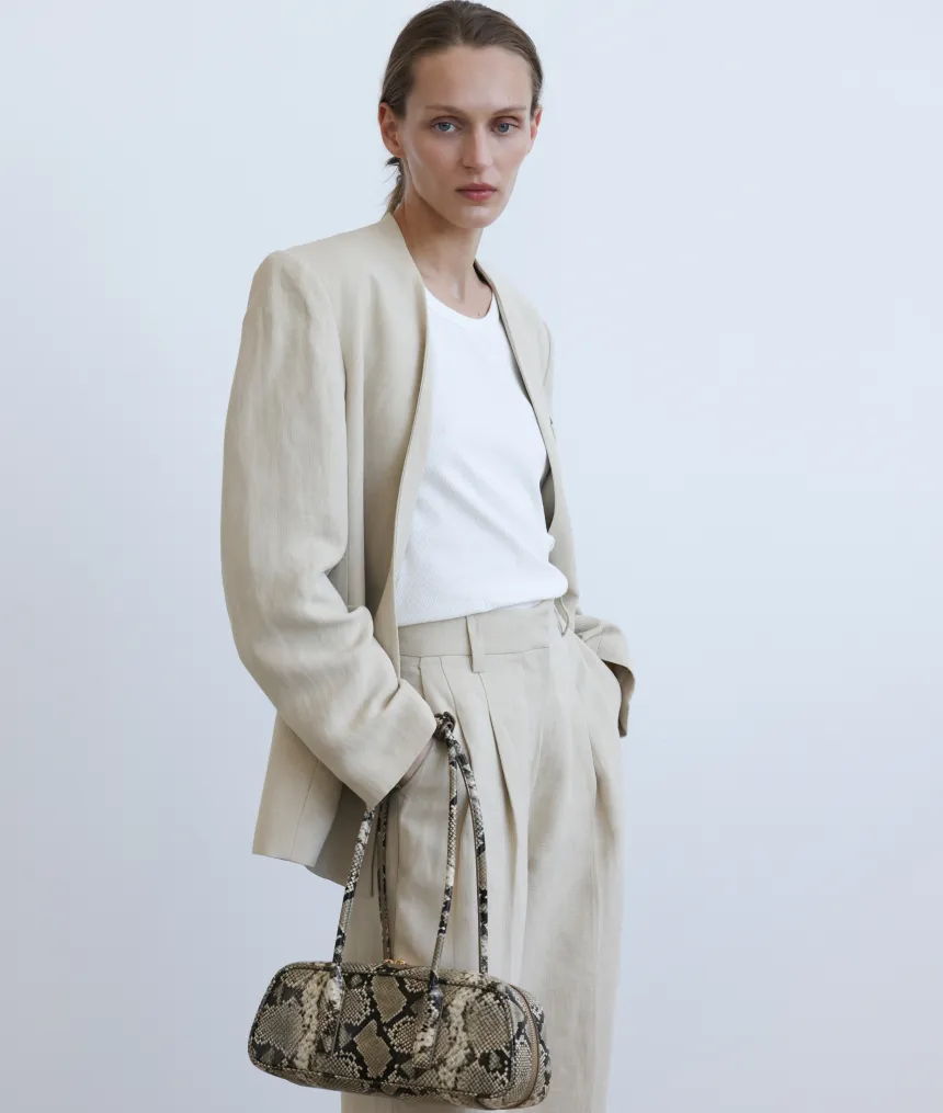 A model wearing a beige suit and holding a snake-print handbag.
