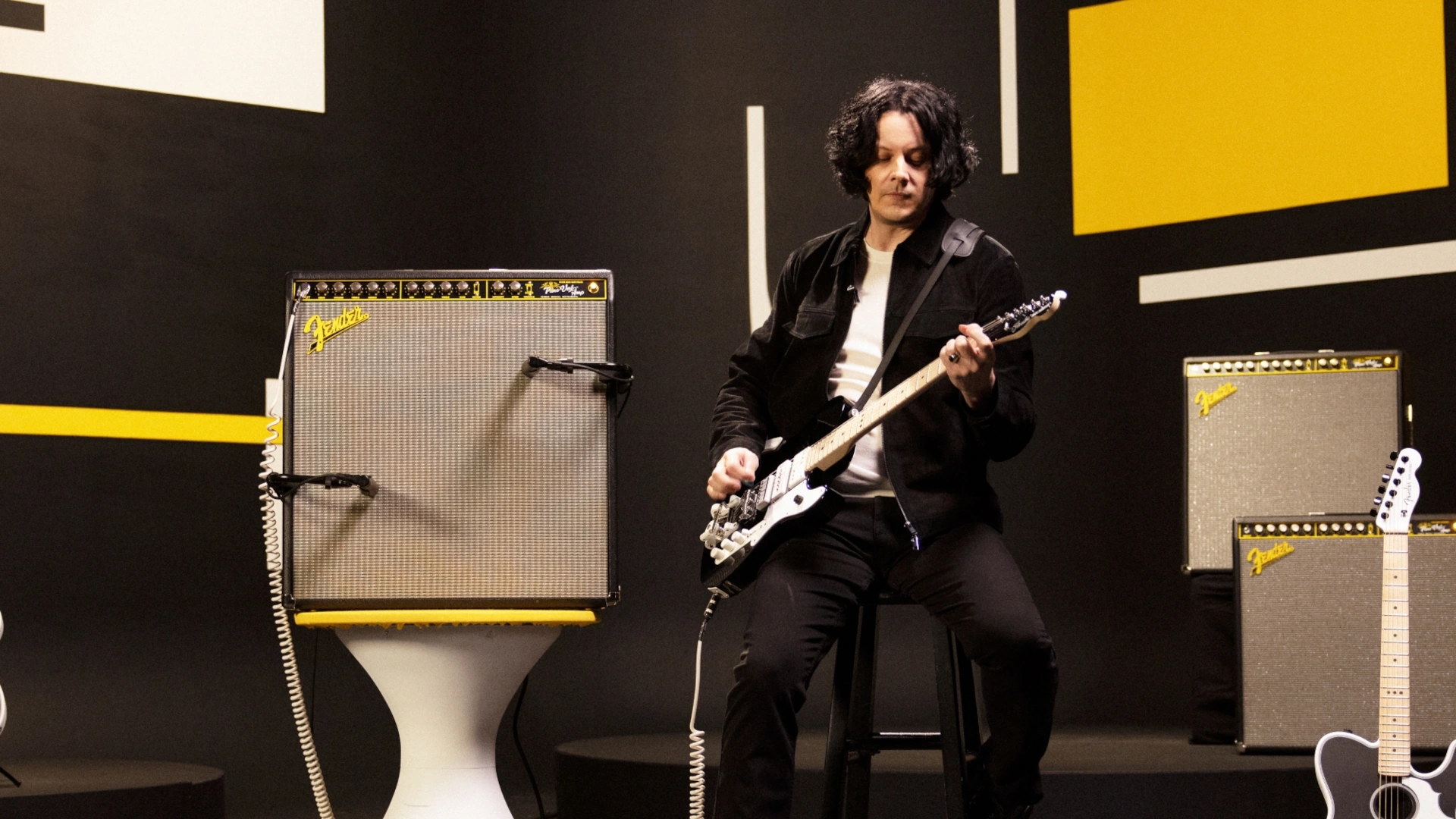 Jack White playing his signature Triplecaster guitar next to amplifiers in a modern setting.