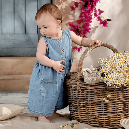 Baby in a denim dress standing next to a wicker basket with flowers.