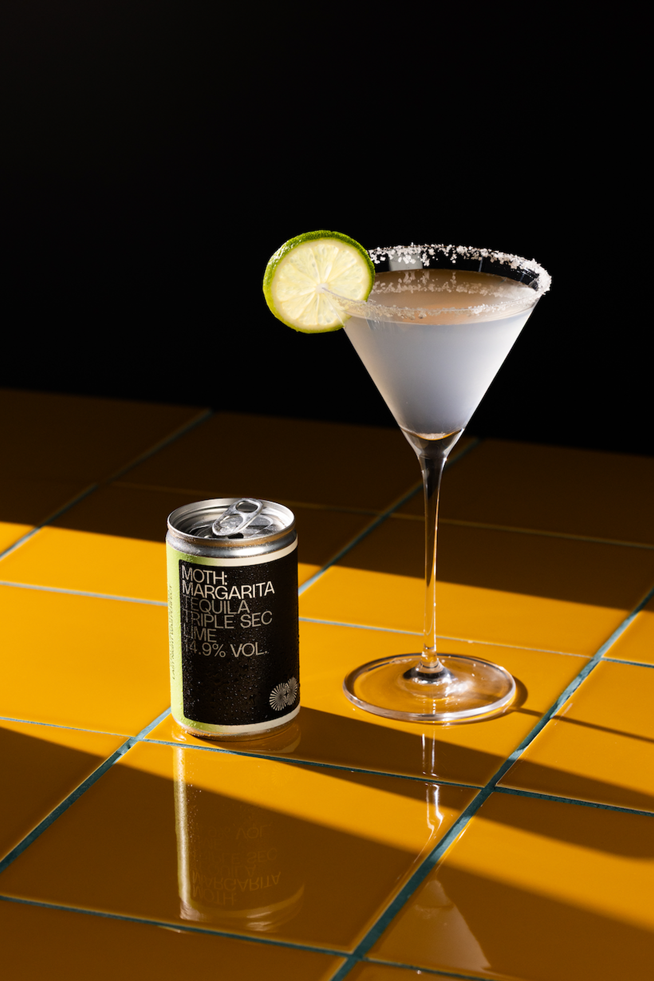 A cocktail with a lime slice next to a can on a yellow surface.