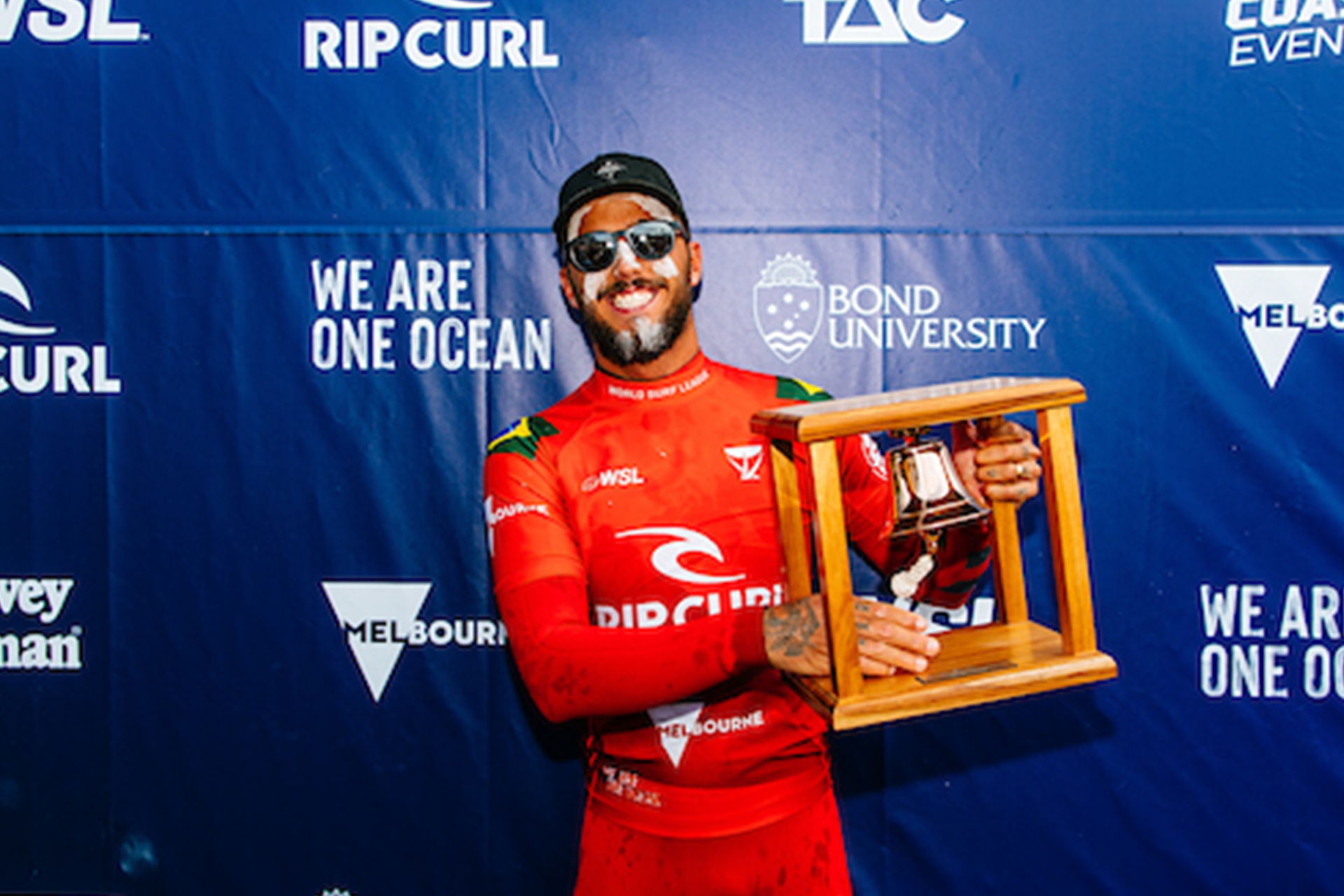 Person in red wetsuit holding a trophy, smiling against a blue backdrop.