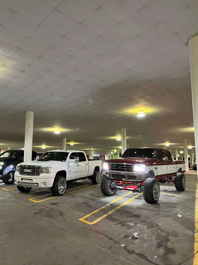 Cade's white lifted 2012 GMC Sierra 2500HD parked beside a lifted two-tone Ford F-Series pickup in an indoor parking garage Nightmare Edition install showcase photo 1
