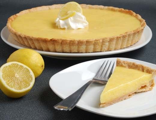 Lemon tart with a slice, whipped cream, and fresh lemons on the side.