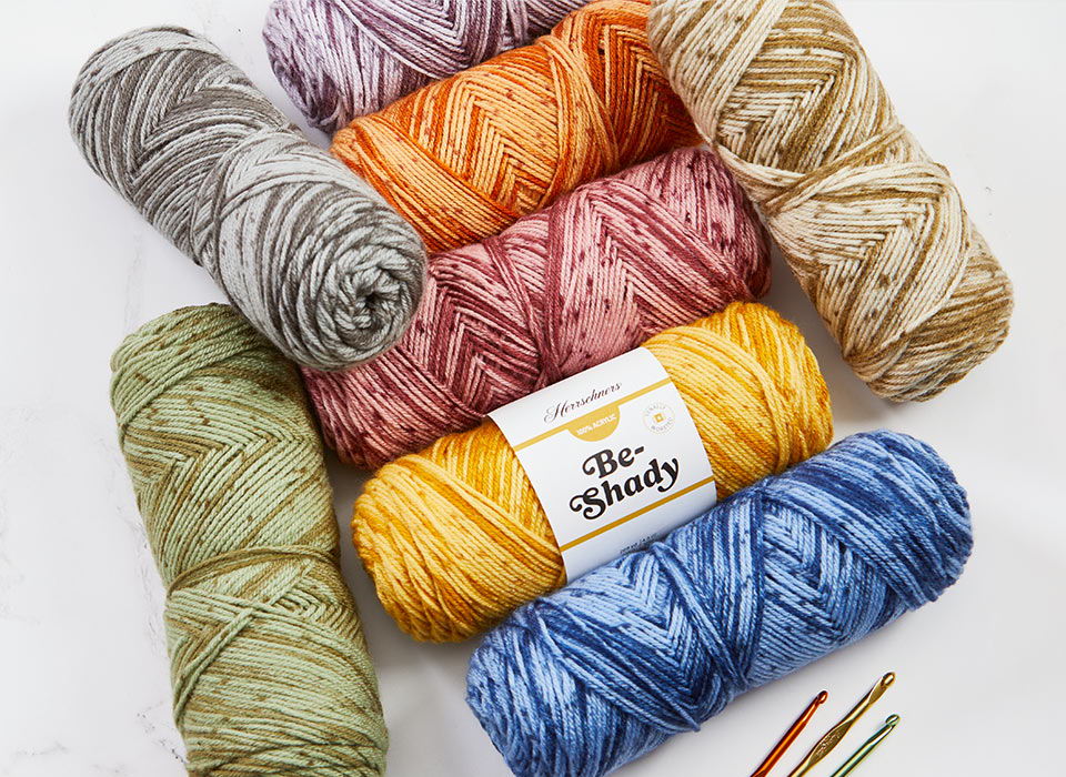 A collection of colorful Herrschners Be-Shady yarn balls and crochet hooks on a white background.