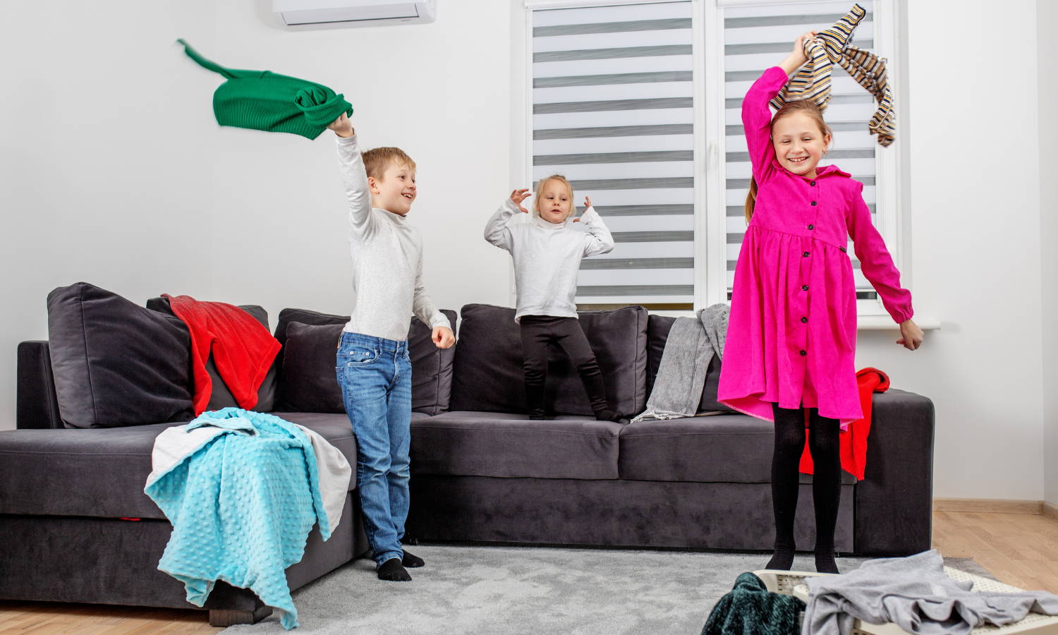 kids in a living room on a sofa throwing clothes around and making a mess