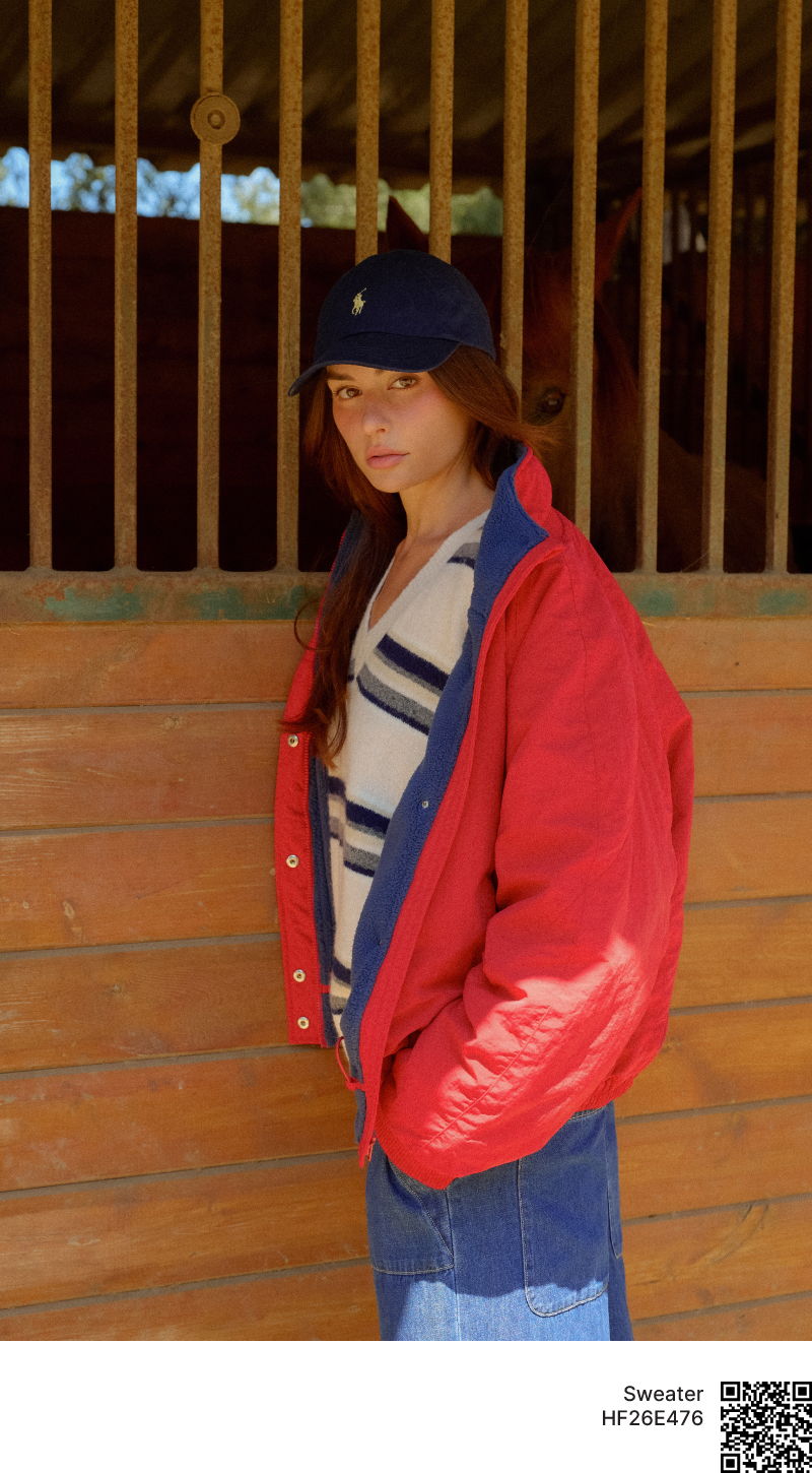 Young woman in a red jacket and blue jeans standing by a barn.