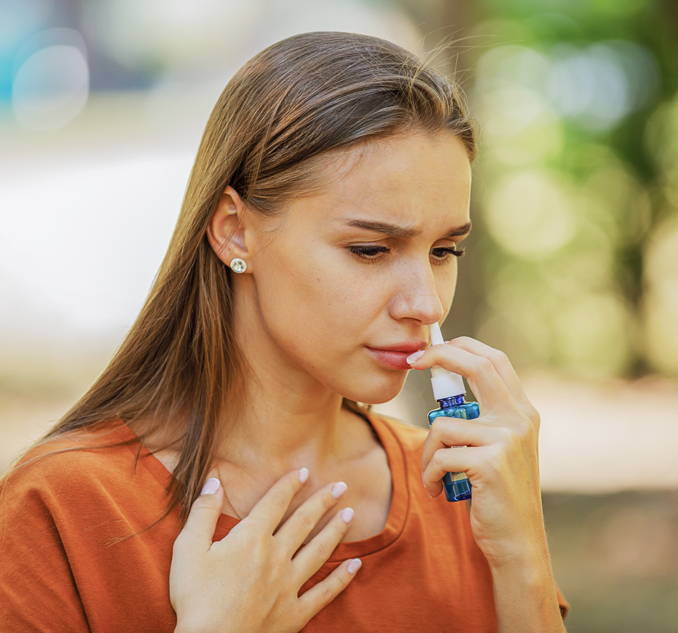 Junge Frau im Freien an einem Sommertag verwendet ein Kortikoid-Nasenspray – dies ist eine verbreitete, kurzfristige Heuschnupfen-Behandlung