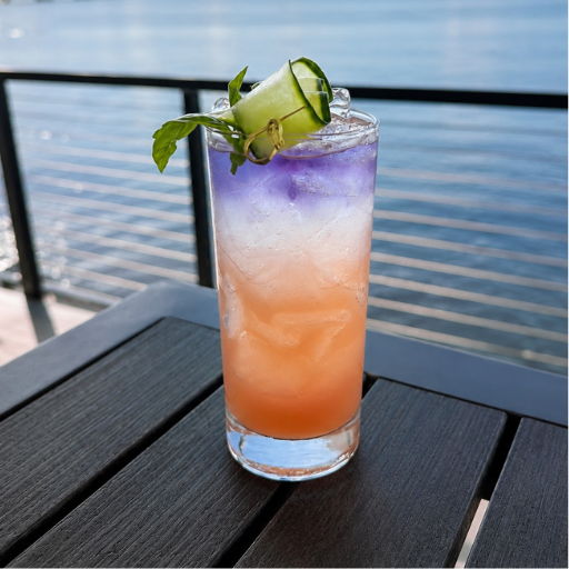 A colorful layered cocktail with cucumber and mint, set by the water.