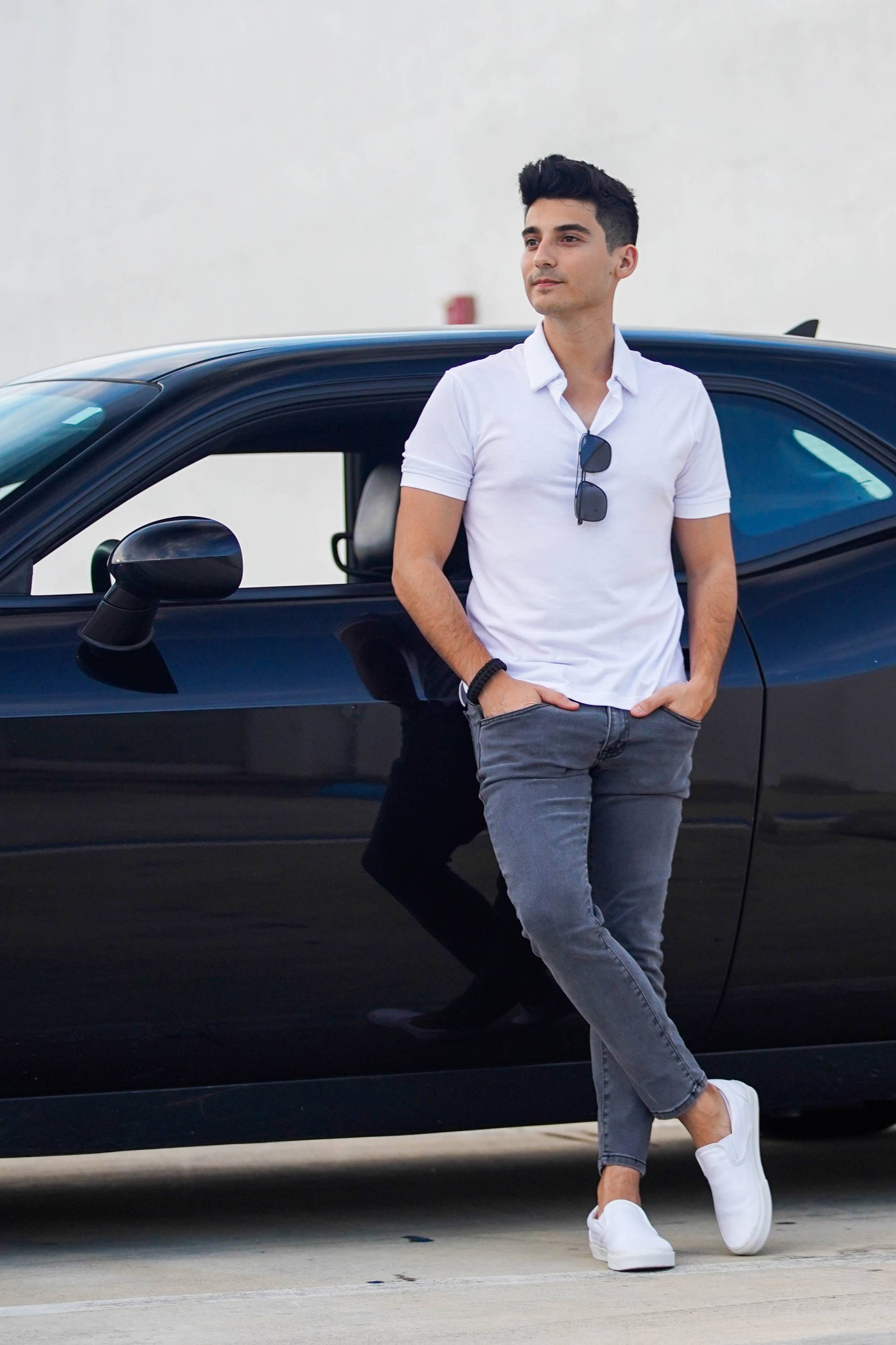 Man leaning against black car wearing white polo shirt, white shoes, and hands tucked into gray jeans pockets from under510.com