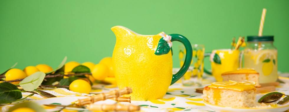 Eine festliche und lebhafte Sommertischdekoration mit einem strukturierten Krug in Zitronenform, Tassen mit Zitronenmotiven und einem Glas mit Zitronenscheiben und einem Strohhalm. Der Tisch ist mit einer Tischdecke mit Zitronendruck, frischen Zitronen und einem Stück Zitronenkuchen vor einem leuchtend grünen Hintergrund geschmückt.