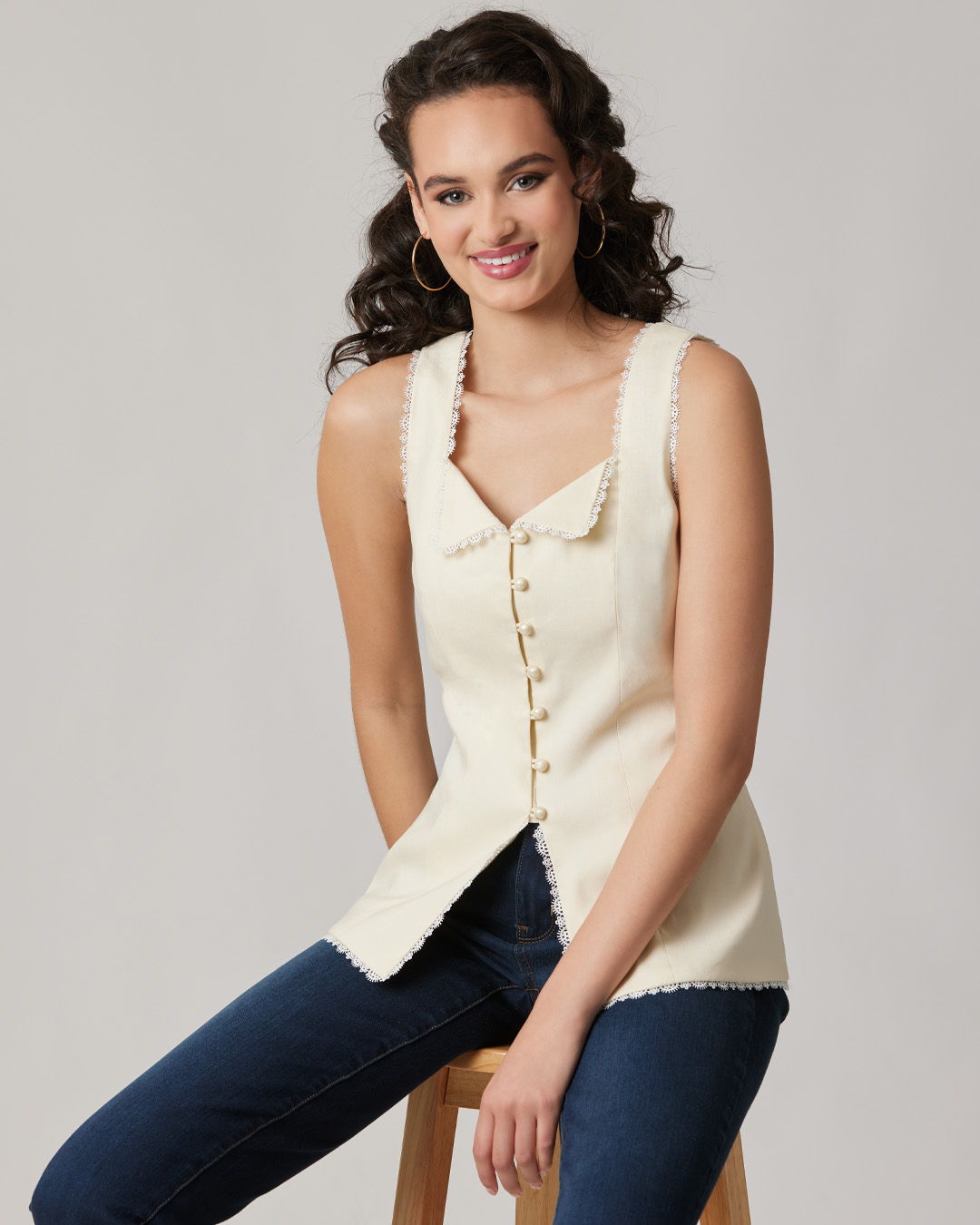 A woman in a cream top and jeans sitting on a stool.