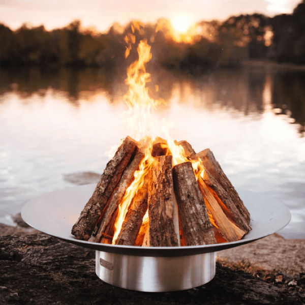 Stainless steel wood-burning fire pit with stacked logs beside a lake at sunset