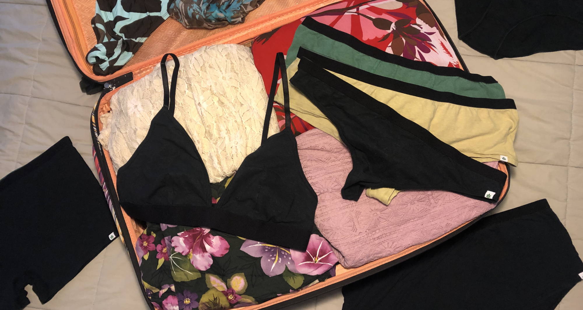 suitcase open on the ground with multicolor clothes folded inside and a black bra and thong underwear laying flat on top
