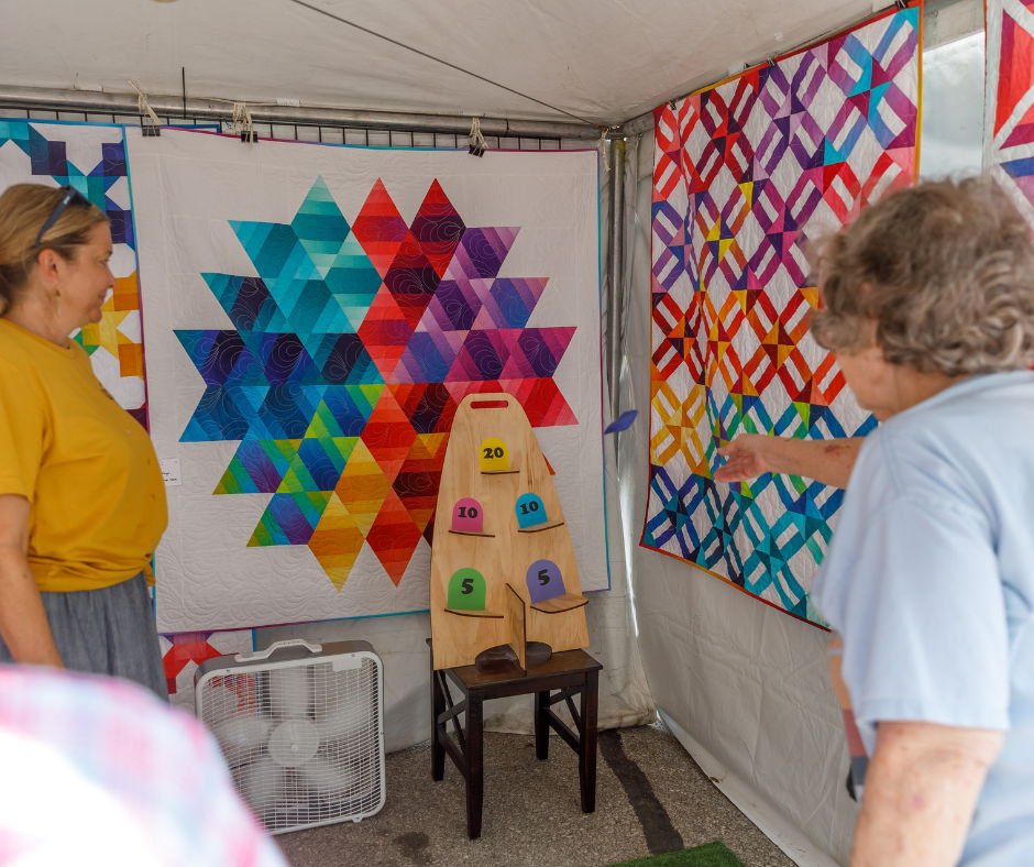 Two women discussing colorful quilt patterns in a tent display.