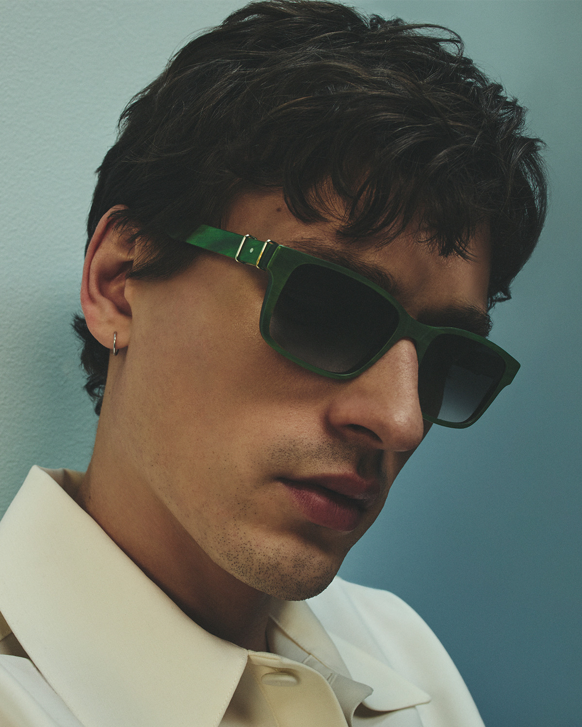 Young man wearing sunglasses, with tousled hair, against a blue background.