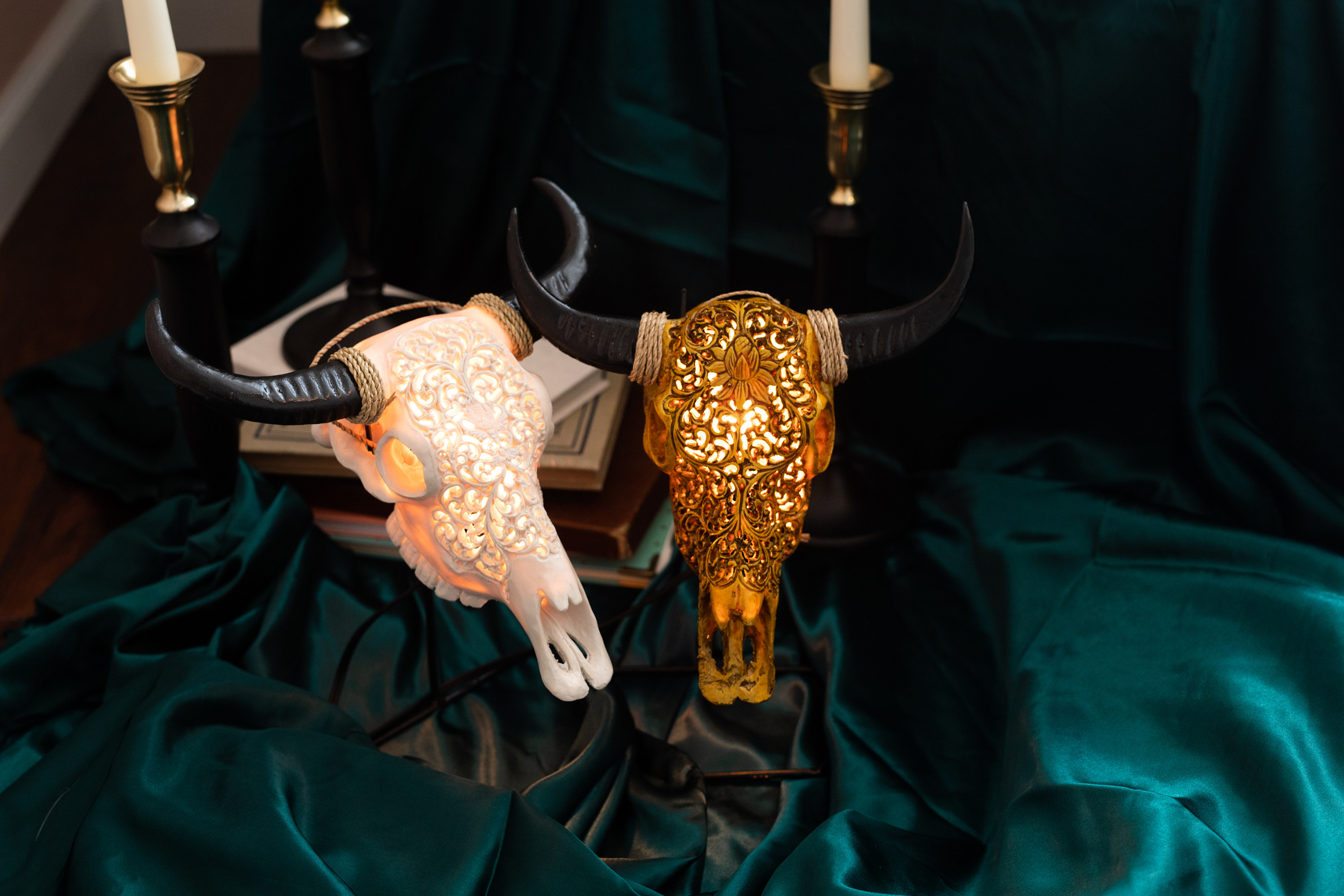 Two hand-carved skull lamps are standing on a green velvet cloth with books and candles in the background.