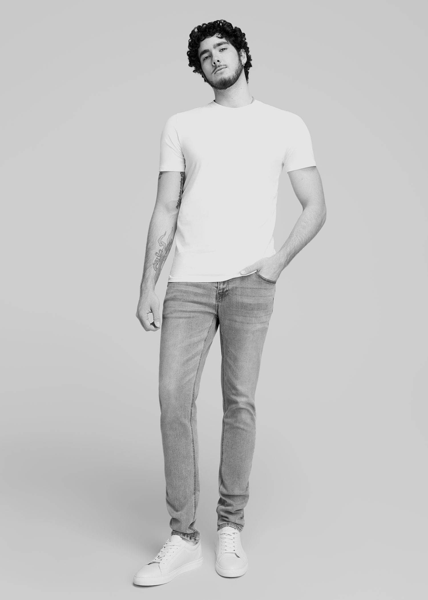 Tall man standing with his hand in his pocket wearing a white tshirt and light jeans