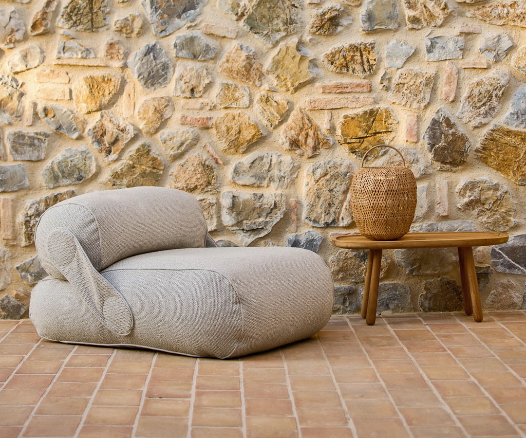 A cozy Cane-line outdoor chair next to a wooden table against a stone wall.