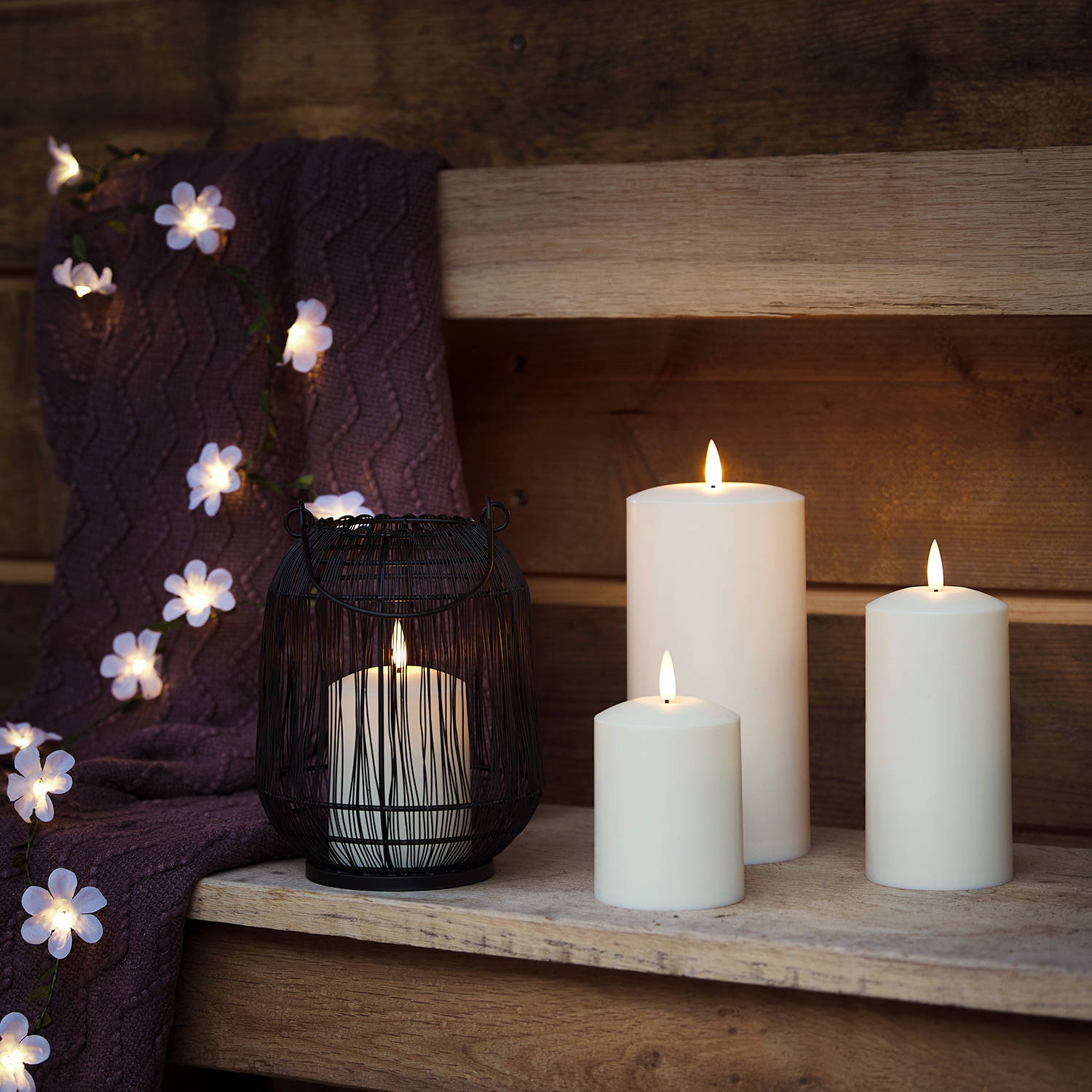 3 chapel candles and a black lantern on a table with a white flower garden in the background.